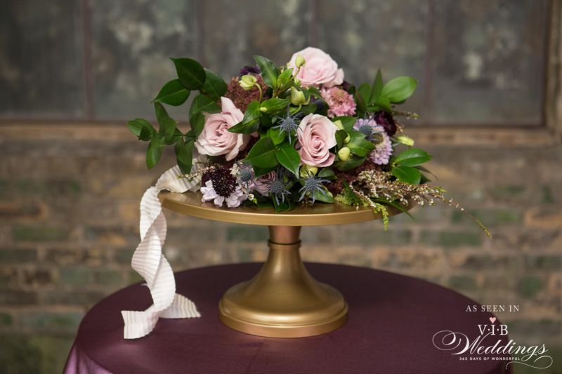 A gold cake stand filled with flowers is sitting on a purple table.