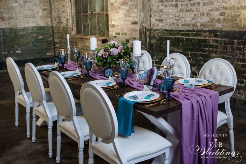 A long table with white chairs and purple and blue decorations.