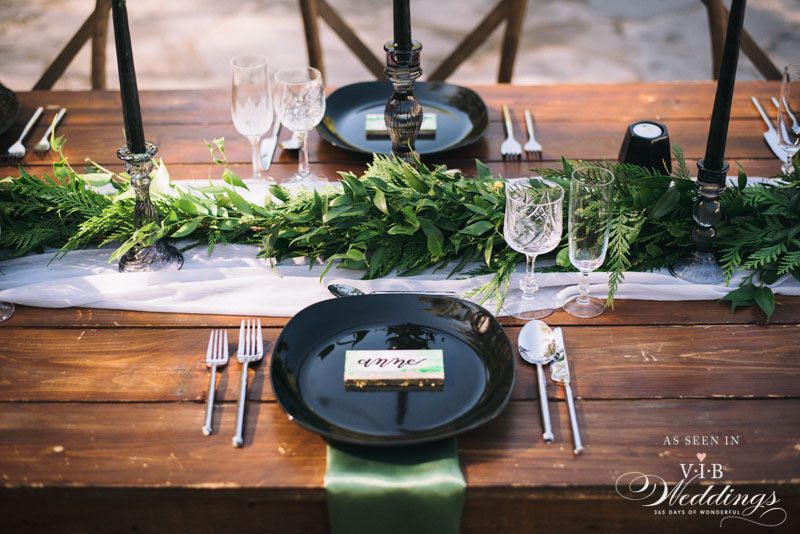 A wooden table with black plates , silverware , and a green table runner.