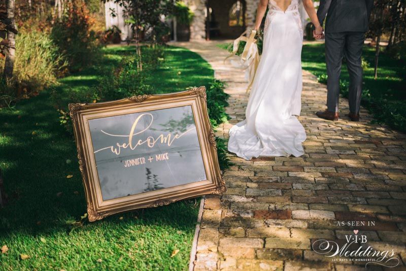 A bride and groom are walking down a brick path next to a welcome sign.