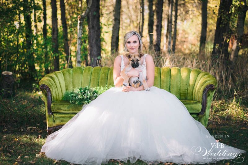 A bride in a wedding dress is sitting on a green couch in the woods holding a dog.