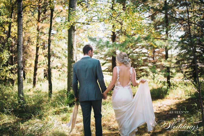 A bride and groom are walking through the woods holding hands.