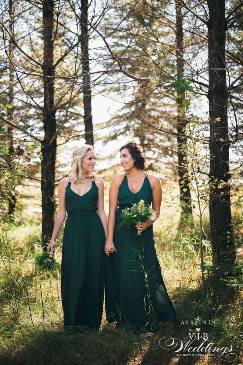 Two bridesmaids in green dresses are standing in the woods holding hands.