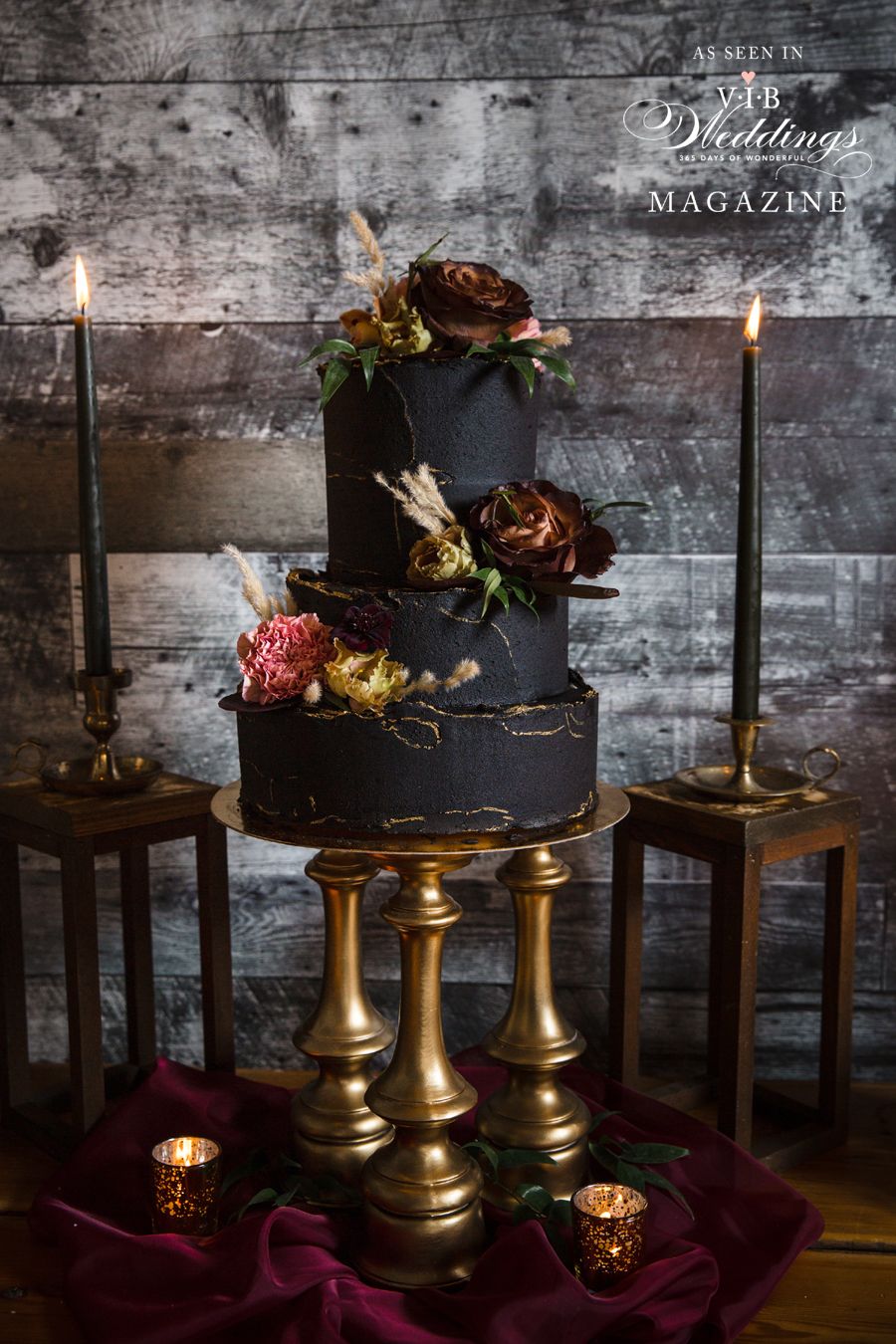 A black wedding cake is sitting on top of a table next to candles.