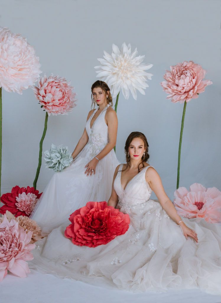 Two women in wedding dresses are sitting next to each other in front of giant paper flowers.