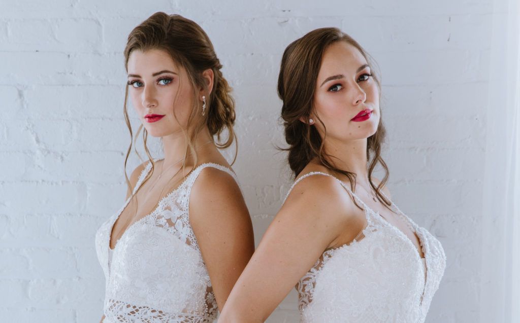 Two women in wedding dresses are standing back to back against a white wall.