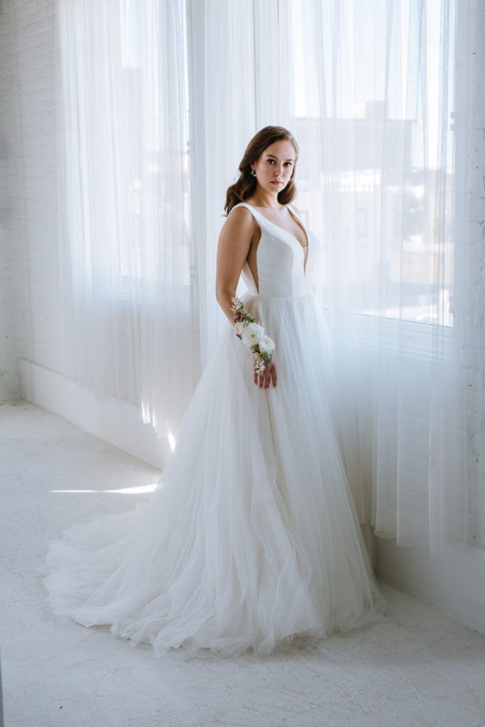 A bride in a wedding dress is standing in front of a window holding a bouquet of flowers.