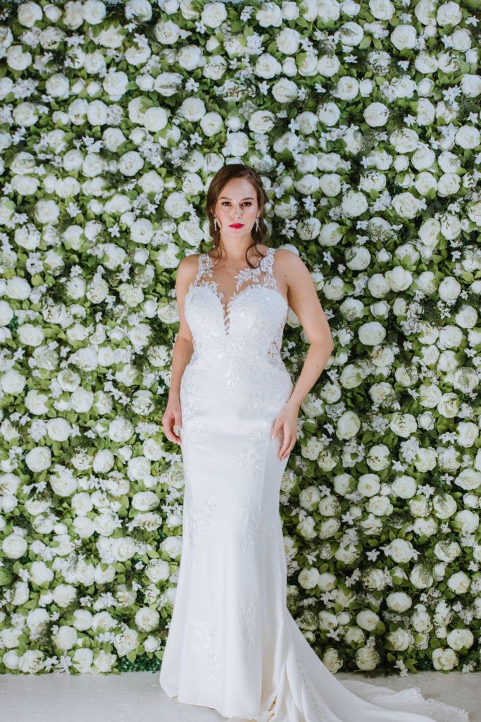 A woman in a wedding dress is standing in front of a wall of white flowers.