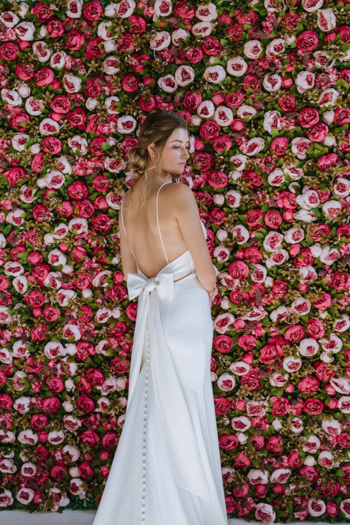 A woman in a wedding dress is standing in front of a wall of flowers.