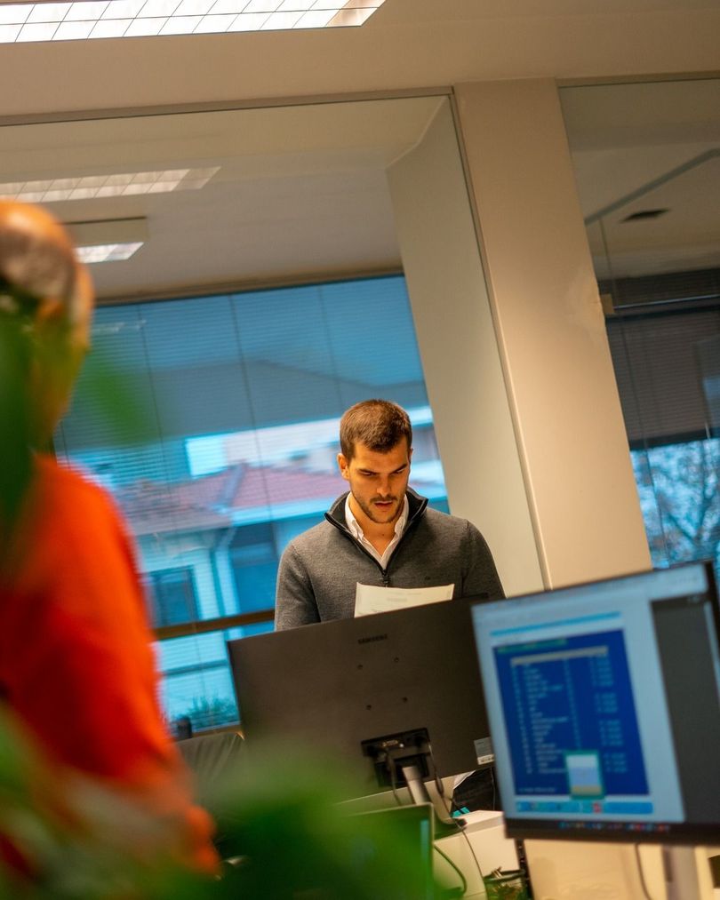 Homem em pé diante de uma mesa de escritório, olhando para um monitor em um escritório iluminado com grandes janelas.