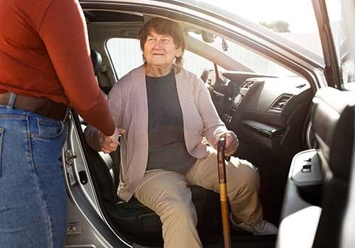 Woman seated in a car doorway, holding a cane, with another person helping her get in