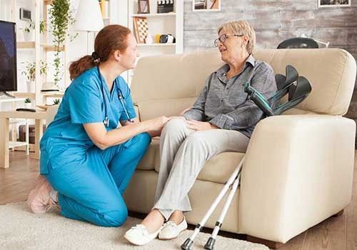 Caregiver in blue scrubs talks with an elderly patient on a couch beside crutches in a bright living room