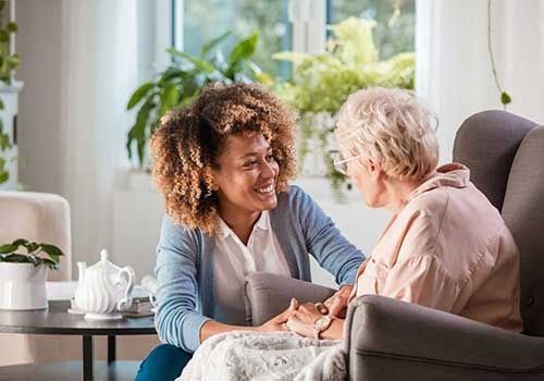 Caregiver smiling and holding hands with an older woman in a bright living room