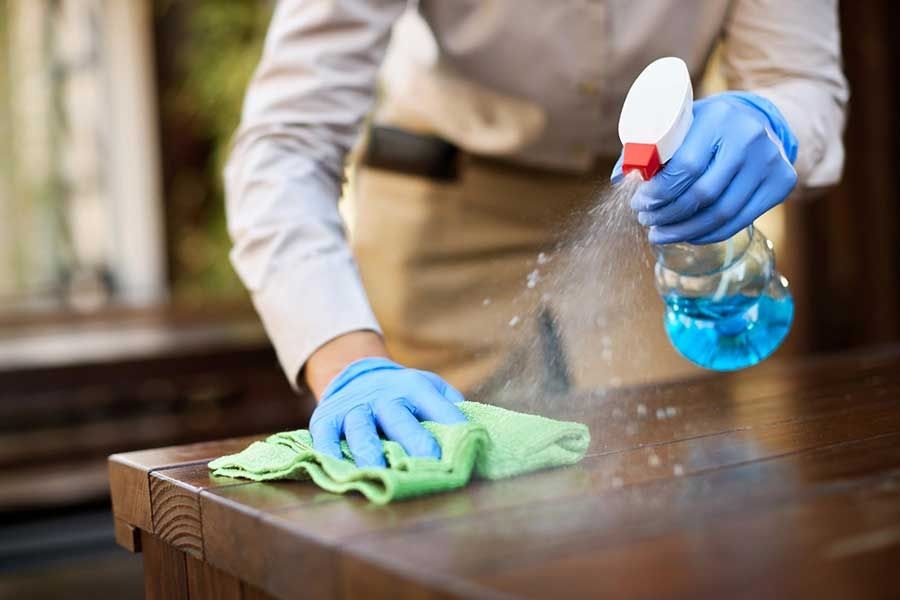 Blue-gloved hands cleaning a wooden table with spray and green cloth