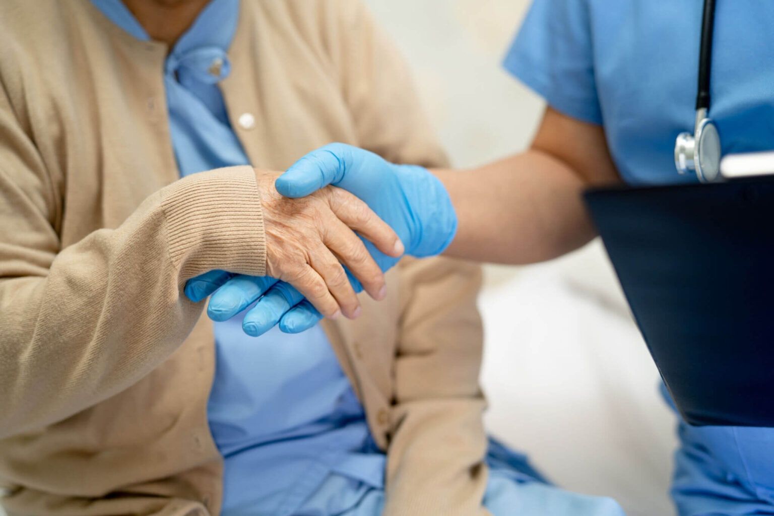 Caregiver in blue gloves gently holds an elderly patient’s hand in a hospital setting