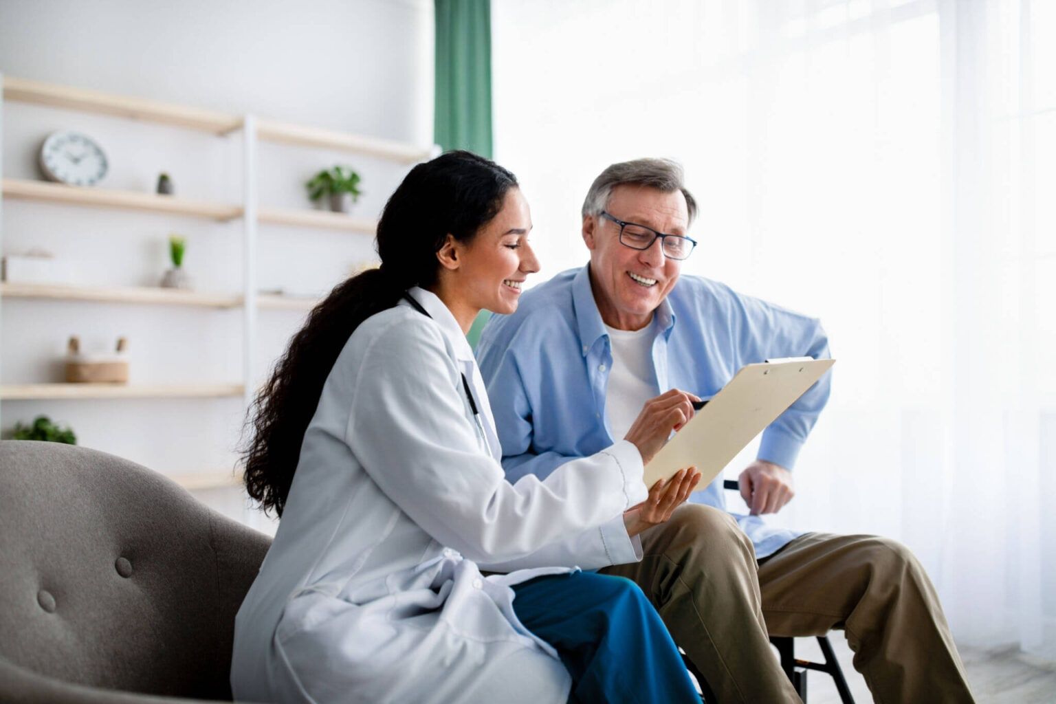 Two people in medical coats reviewing a clipboard on a couch in a bright living room.