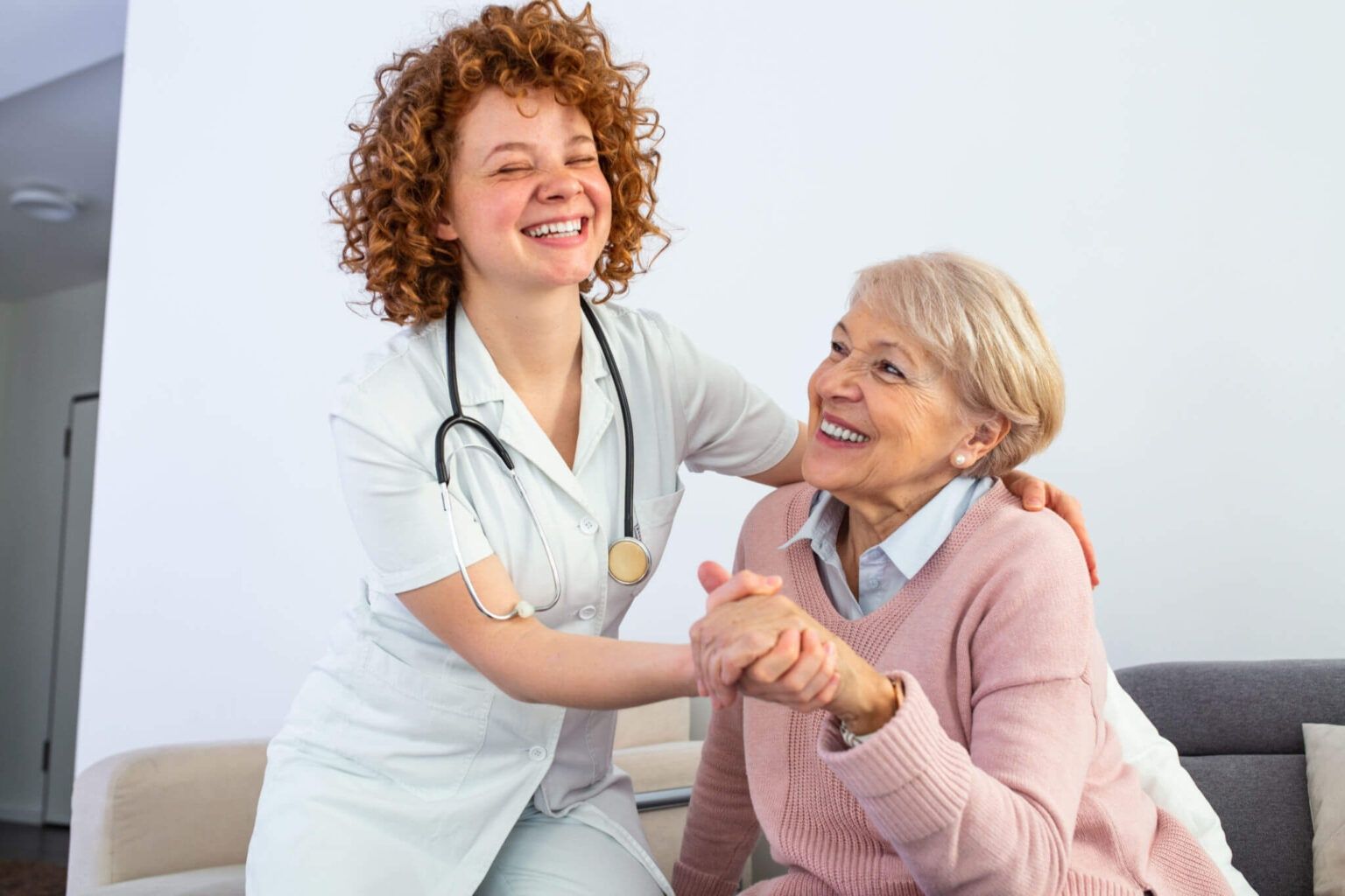 Caregiver in white scrubs smiles while holding hands with a smiling elderly woman on a couch