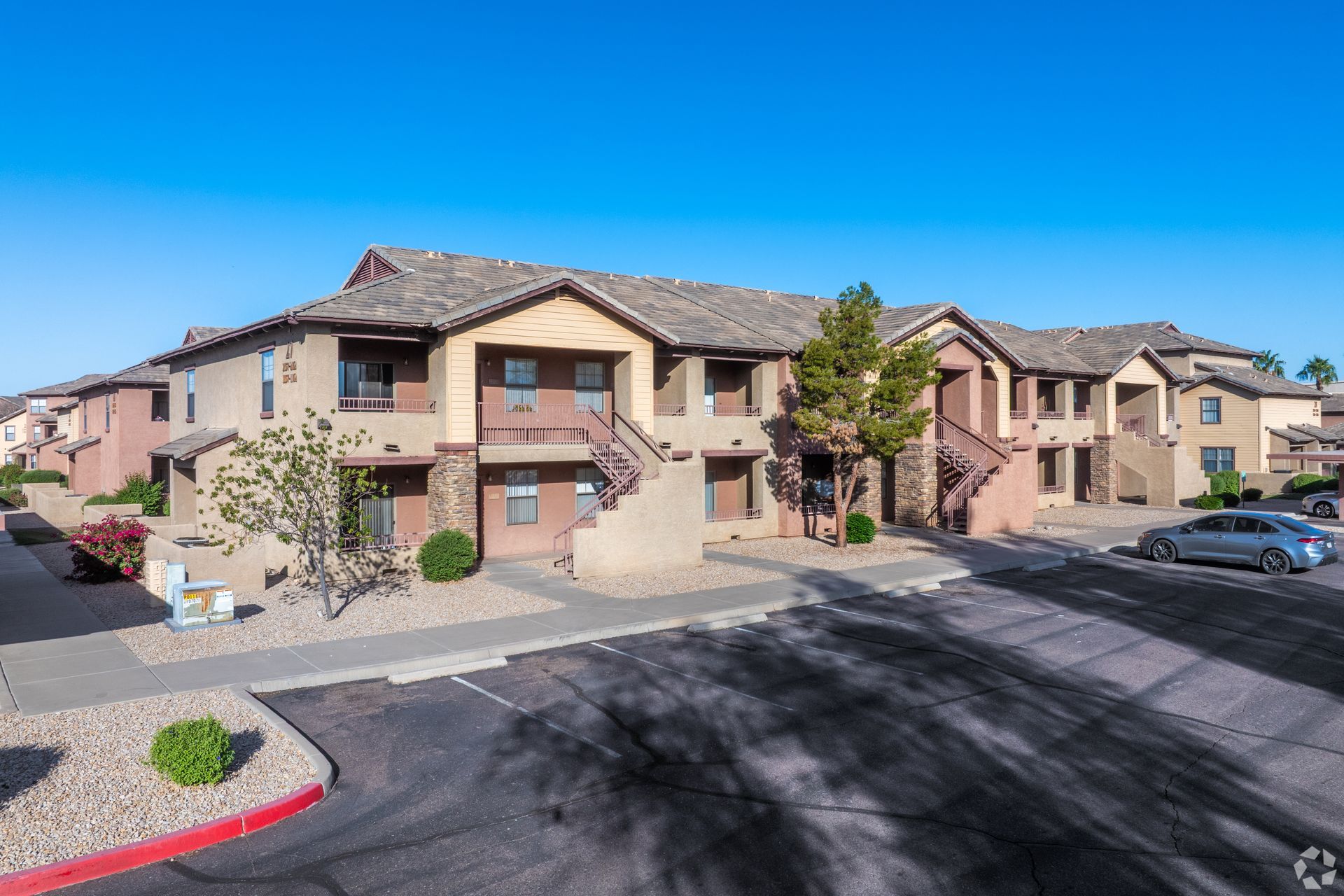 A multi-story apartment building with tan stucco walls and a tile roof, featuring a parking lot under a clear blue sky.