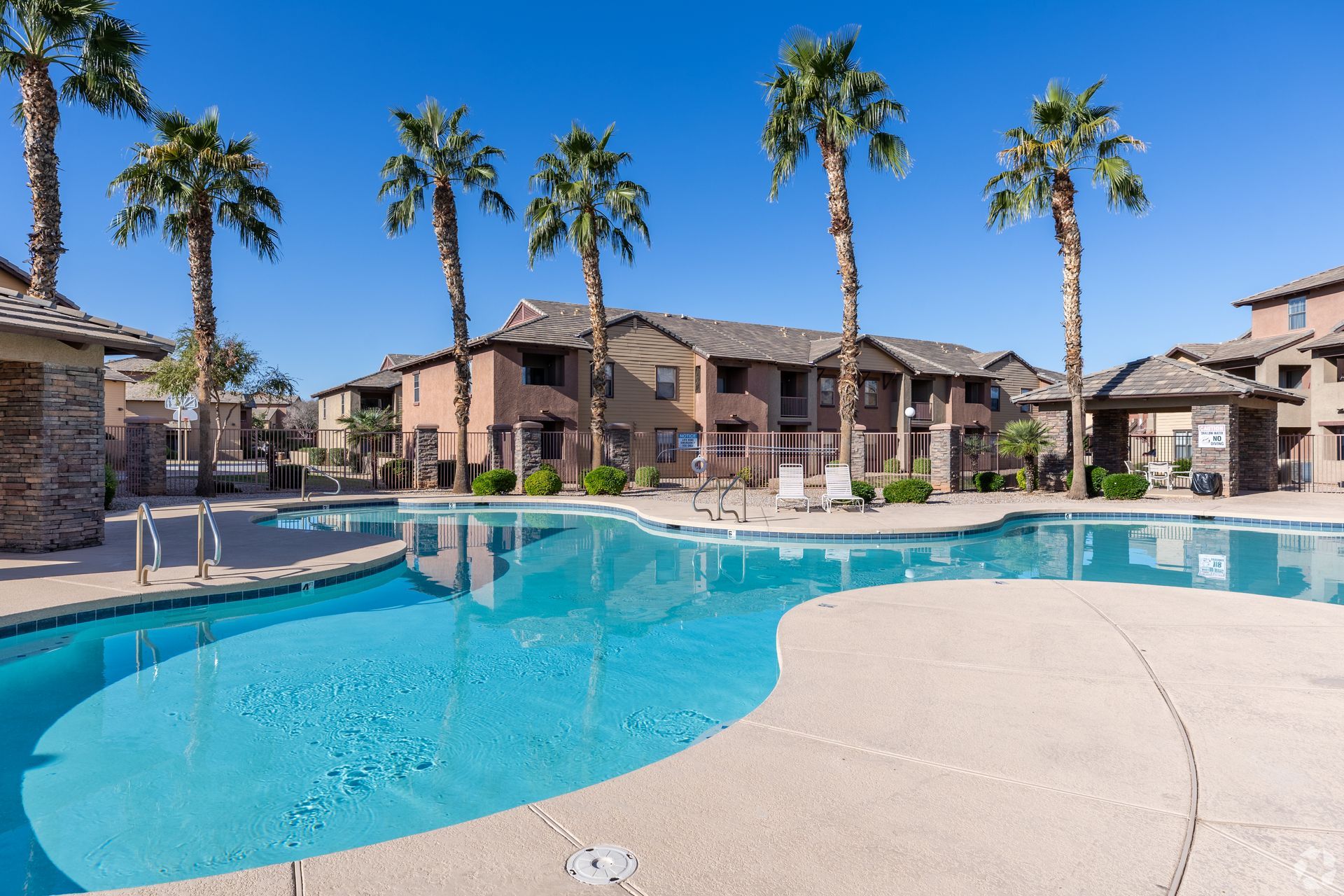 A resort-style swimming pool at an apartment complex, featuring palm trees and multi-story tan buildings under a blue sky.