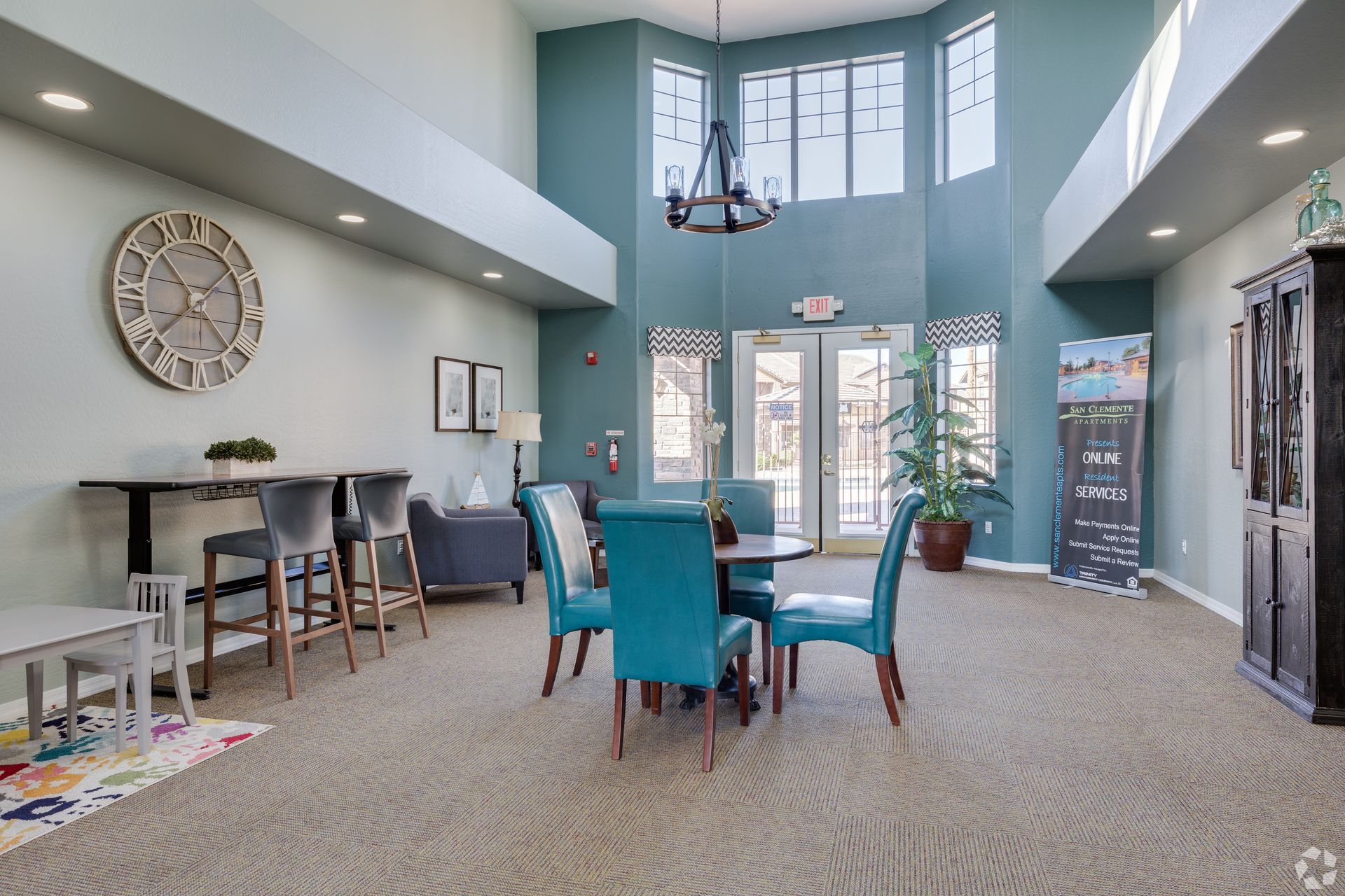 A brightly lit, modern community room featuring a tall bar table with chairs, a round dining table, and large windows.