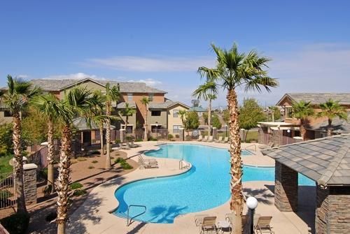 An outdoor swimming pool surrounded by a patio, palm trees, and multi-story apartment buildings on a sunny day.