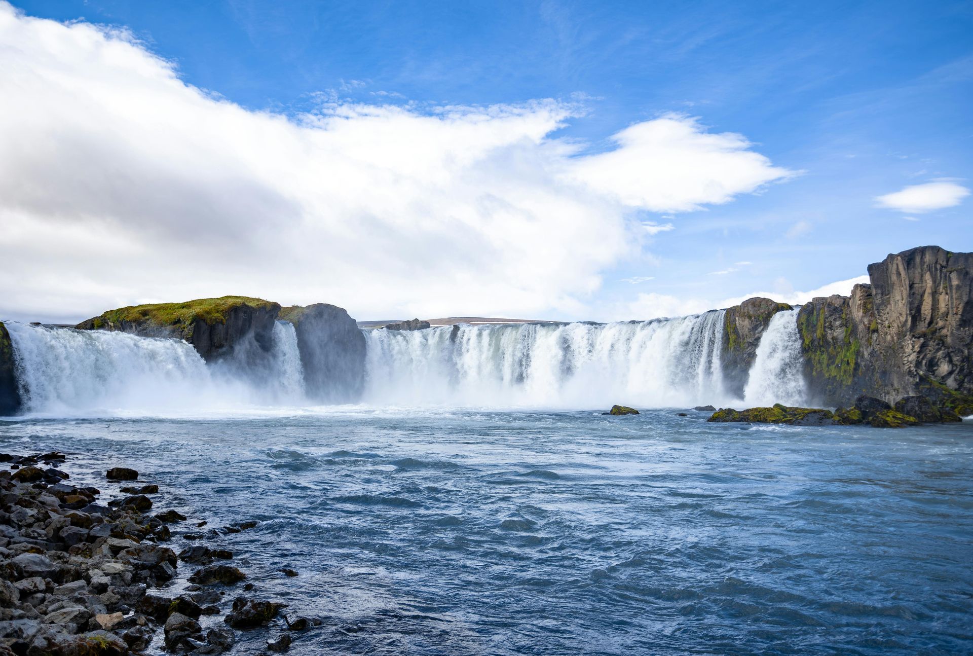 Wide waterfall cascading into a rocky blue river under a cloudy sky