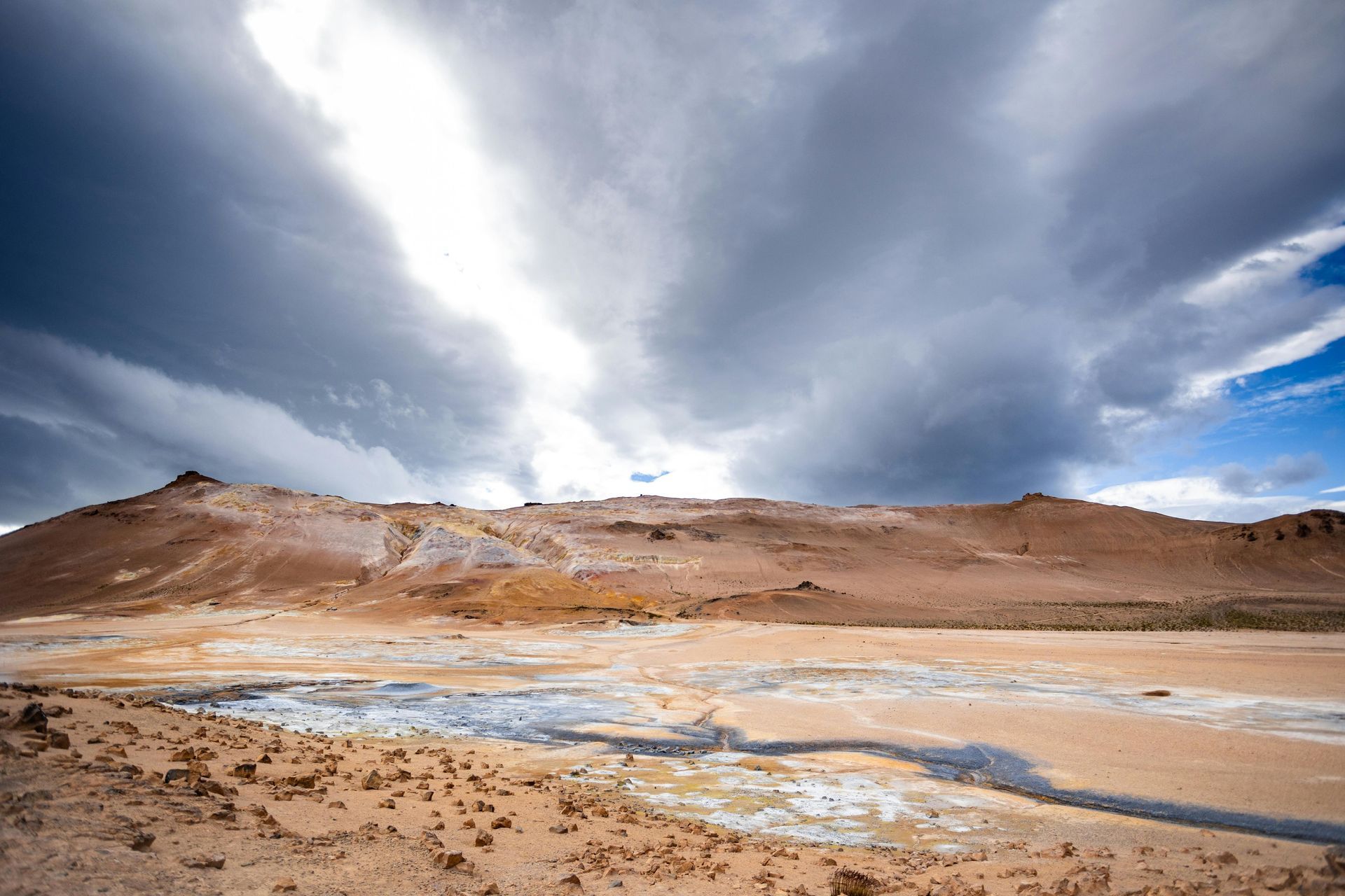 Steam rising from a muddy geothermal pool beneath dramatic cloudy sky in a barren desert landscape
