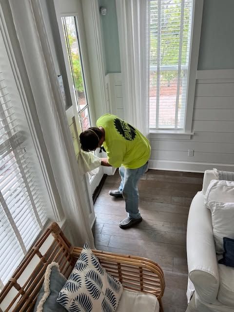Person in a neon shirt bending by a window in a bright room with white walls and wicker chairs