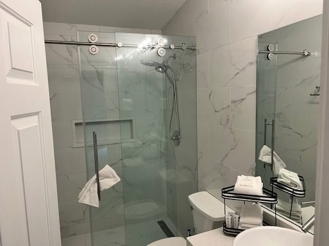 Glass shower enclosure and toilet in a small marble-tiled bathroom with a sink and mirror cabinet