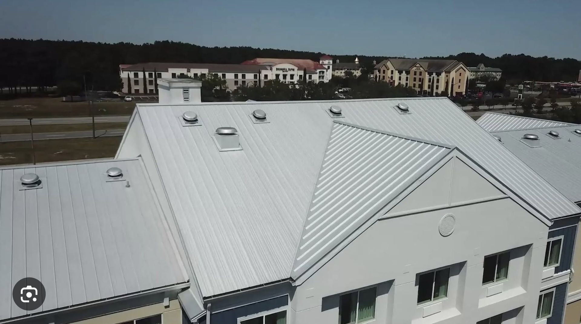 White metal-roofed building complex with skylights and nearby residential houses in the background