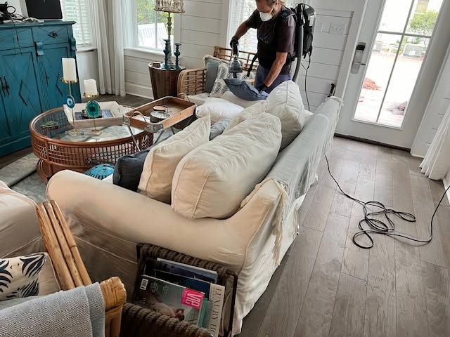 Person vacuuming a white couch in a bright living room with wicker tables and cords on the floor