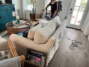 Living room with white sofa, blue cabinet, woven tables, and a person vacuuming near the door