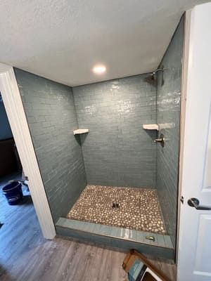 Empty tiled shower stall with blue-gray walls, pebble floor, and built-in corner shelves.