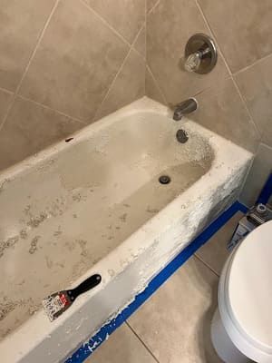 Dirty bathtub in a tiled bathroom with a paintbrush and blue tape along the rim