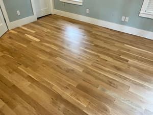 Empty room with glossy hardwood floors, white walls, and a window with blinds.