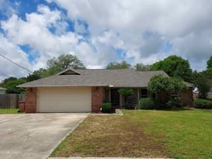 Single-story brick house with two-car garage, driveway, and front lawn under a partly cloudy sky