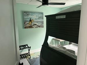 Small mint bedroom with a black canopy bed, beach painting, ceiling fan, and folding chair.