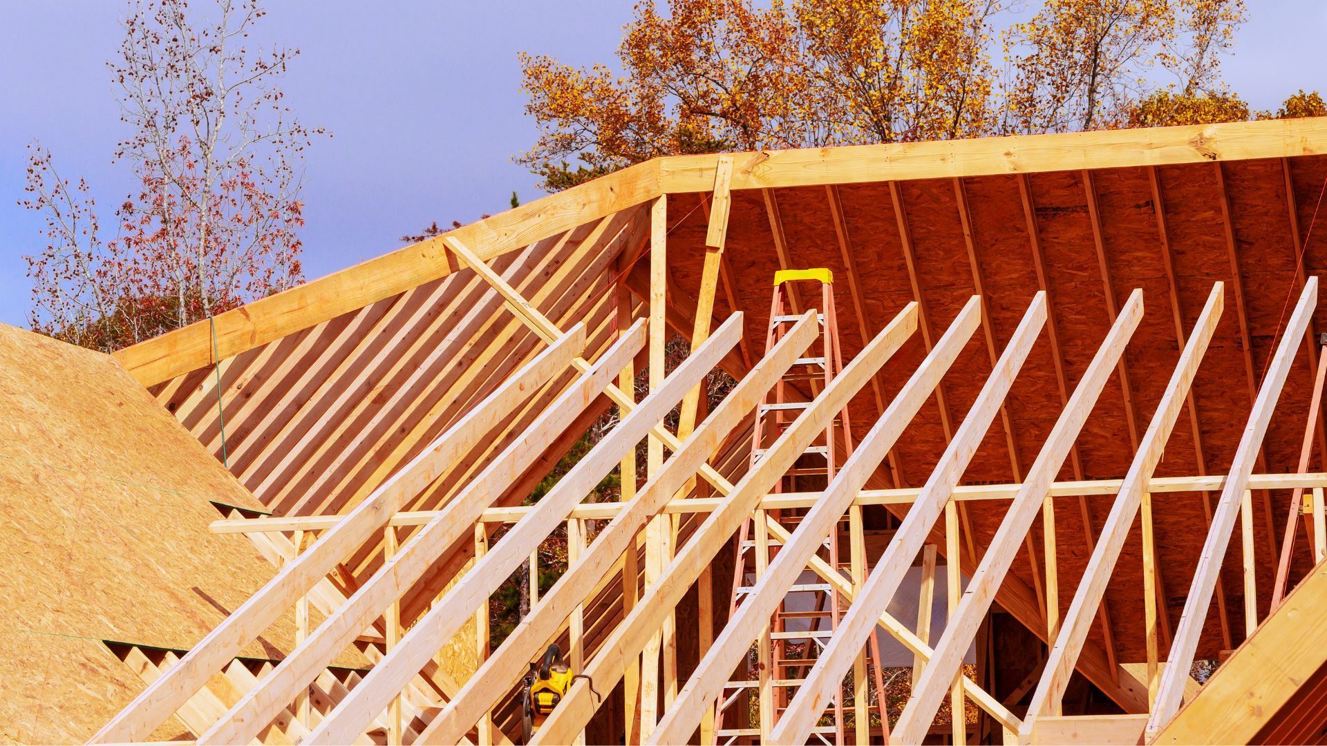Framed house under construction with exposed white rafters and plywood sheathing against a blue sky