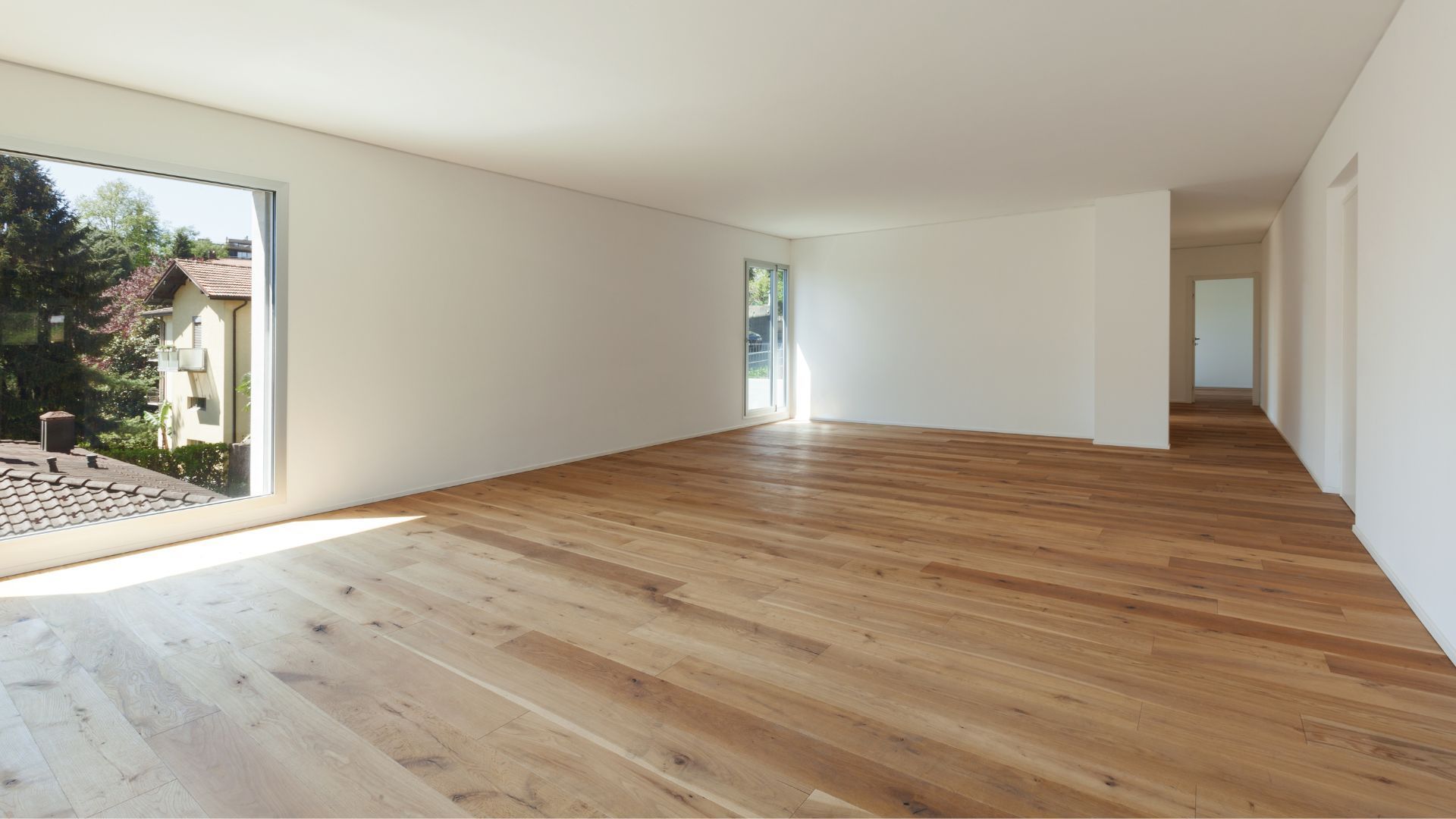 Empty modern living room with light wood floors, white walls, and large windows letting in natural light
