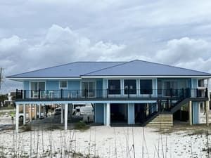 Stilt house on a sandy beach under a cloudy sky, with a blue roof and raised porch.