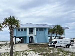 Blue beach house on stilts with palm trees and a white van parked outside under cloudy skies