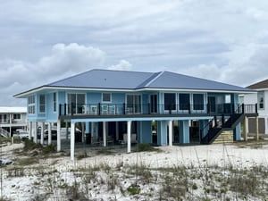 Blue beach house on stilts with covered porch, set among sand dunes under a cloudy sky