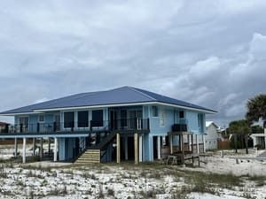 Blue beach house on stilts under a cloudy sky, with sandy dunes and a porch.