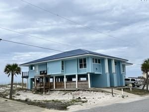 Blue raised beach house on stilts with sandy yard under a cloudy sky