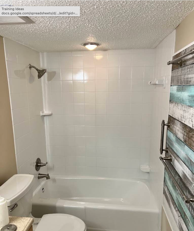 Bathroom with a white tiled shower-tub combo, toilet on the left, and a window with blinds on the right.