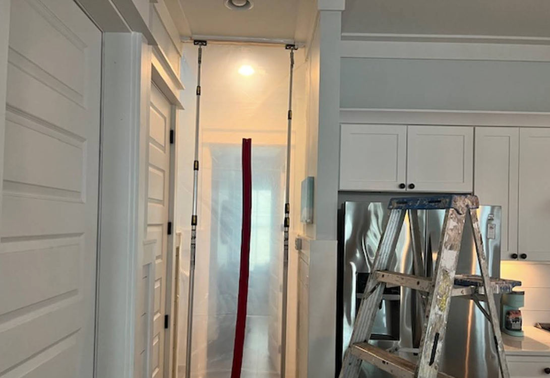 Narrow hallway with hanging plastic sheeting and a ladder in a bright, white kitchen area