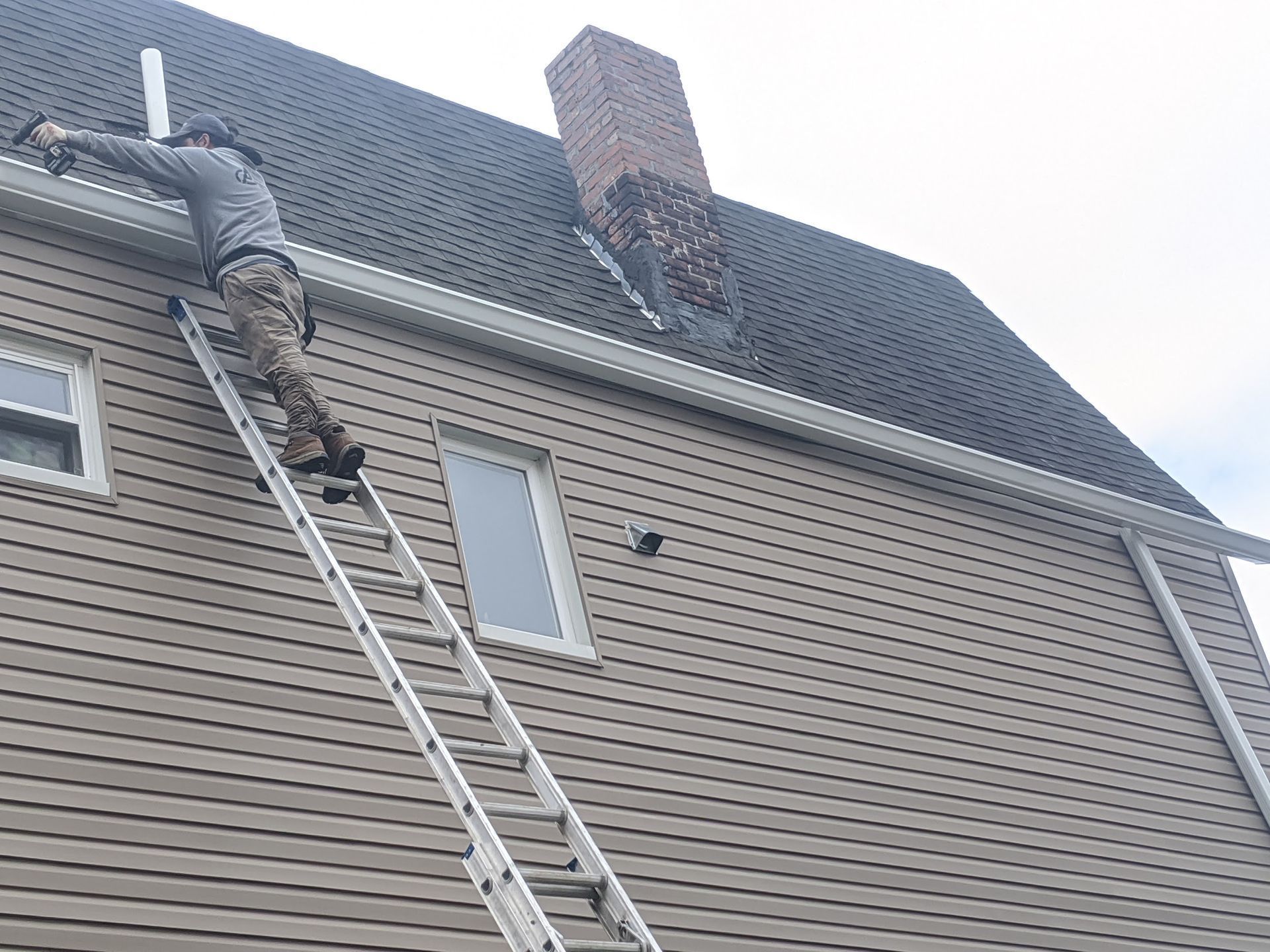 Person on ladder cleaning leaves from roof near chimney.