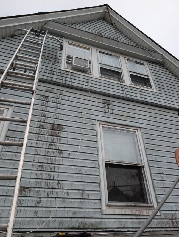 Weathered gray house siding with two windows and a steep gabled roof under overcast sky
