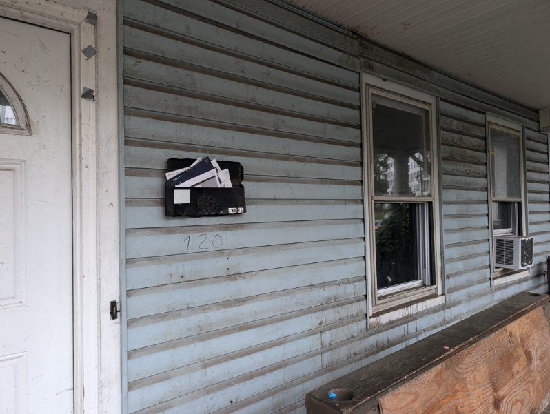 Weathered house siding with mailbox, window, and plywood leaning against the wall