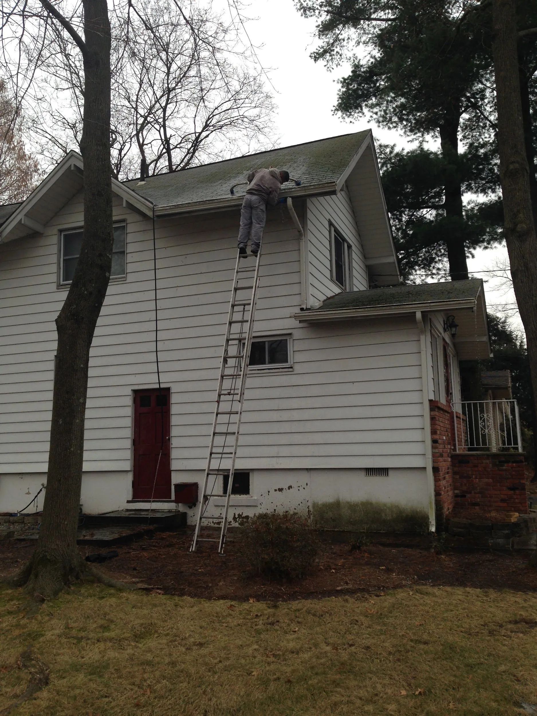 Person on a ladder, working on the roof of a white house. Trees surround the house.